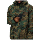 A person wearing a hooded camouflage jacket, holding a black smartphone in one hand. The jacket features a green, brown, and black woodland camo pattern, and the photo is cropped from the chin down against a plain white background.