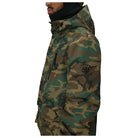 A side profile view of a man wearing a camouflage hooded jacket with a black neck gaiter. The jacket features a green, brown, and tan camo pattern with a black logo on the left sleeve.