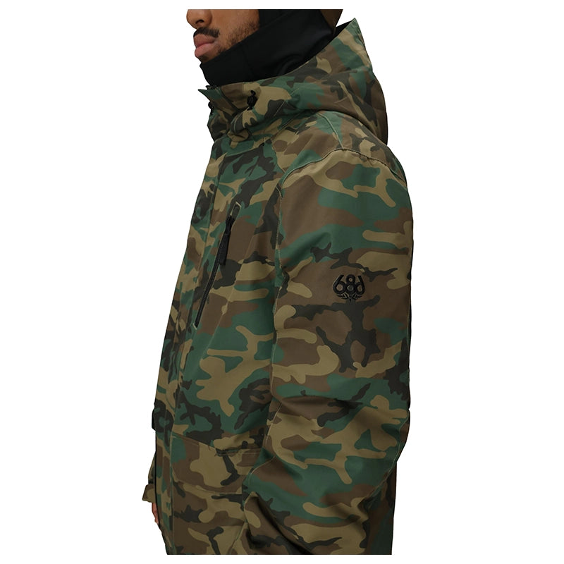 A side profile view of a man wearing a camouflage hooded jacket with a black neck gaiter. The jacket features a green, brown, and tan camo pattern with a black logo on the left sleeve.