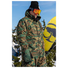A person wearing a green and brown camouflage ski jacket and matching pants, with black goggles and a black beanie, holding a yellow snowboard outdoors against a blue sky and snowy trees.
