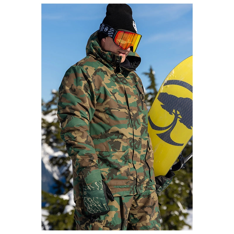 A person wearing a green and brown camouflage ski jacket and matching pants, with black goggles and a black beanie, holding a yellow snowboard outdoors against a blue sky and snowy trees.