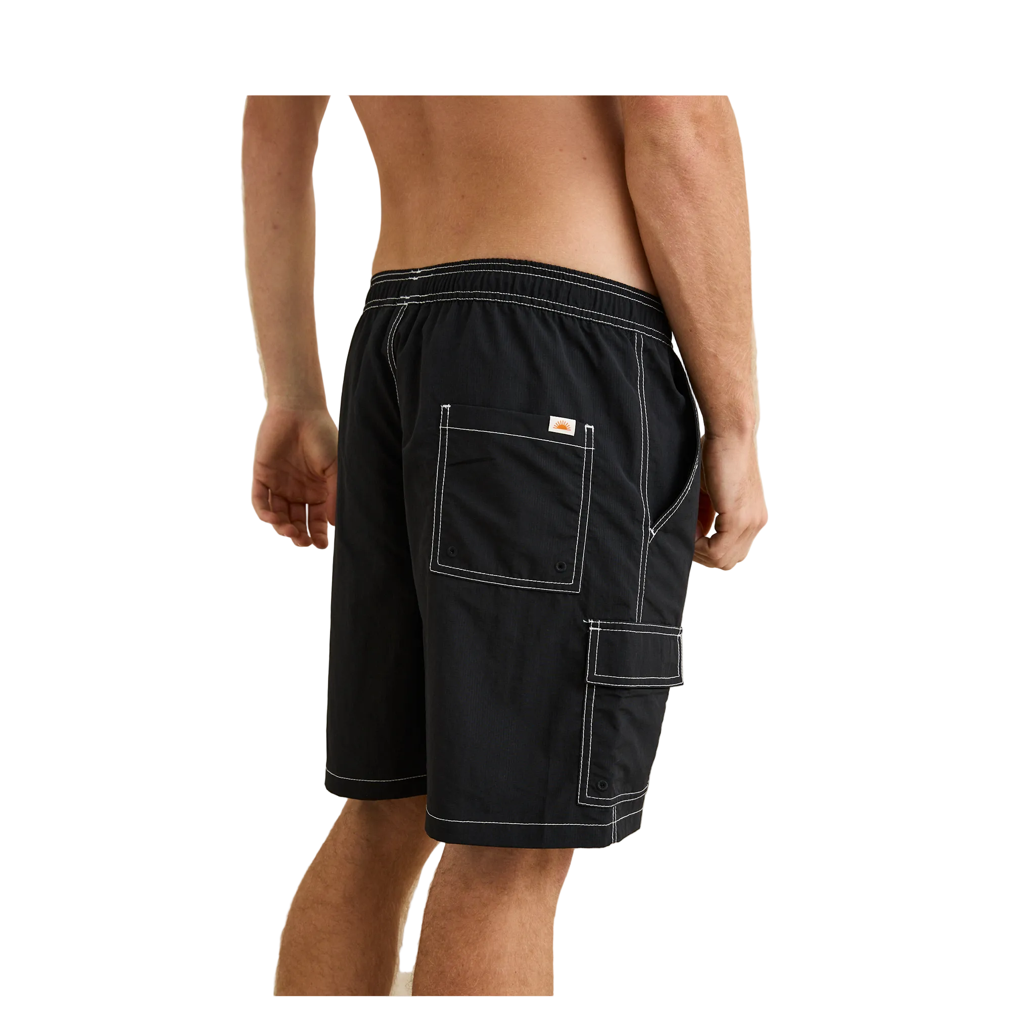 A man wearing black cargo board shorts with white contrast stitching, shown from the back against a light beige background.