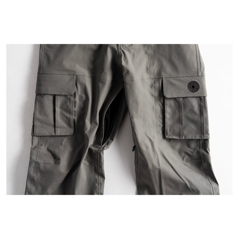 Close-up of dark gray cargo pants showing the right leg with a large flap cargo pocket and a circular black logo patch on the pocket flap, set against a plain white background.