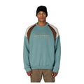 A man wearing a colorblock crewneck sweatshirt by Six Eight Six, featuring a teal body, brown shoulder panels, and cream sleeve panels, paired with a black beanie and khaki pants against a white background.