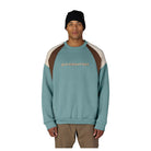 A man wearing a colorblock crewneck sweatshirt by Six Eight Six, featuring a teal body, brown shoulder panels, and cream sleeve panels, paired with a black beanie and khaki pants against a white background.