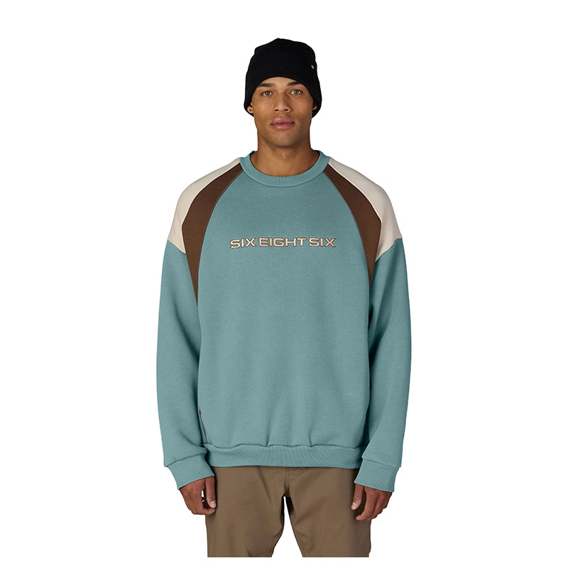 A man wearing a colorblock crewneck sweatshirt by Six Eight Six, featuring a teal body, brown shoulder panels, and cream sleeve panels, paired with a black beanie and khaki pants against a white background.