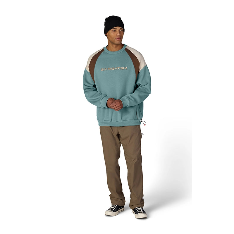 A man modeling a colorblock sweatshirt from Sixty Eighty Four, featuring a teal body, brown shoulder panels, and beige sleeve panels, paired with brown pants, a black beanie, and black sneakers on a white studio background.