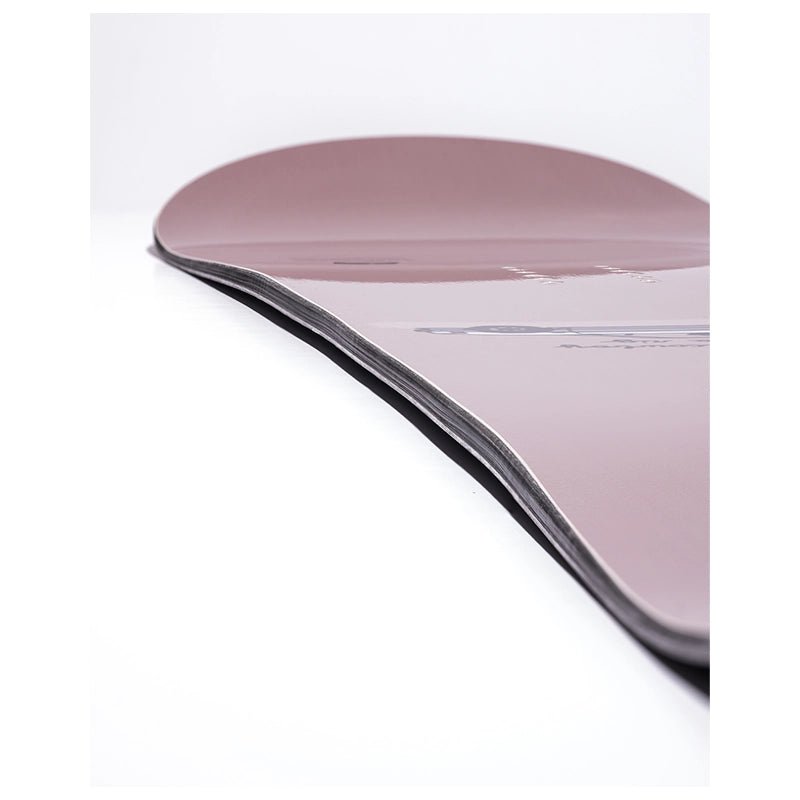 A close-up studio shot of a snowboard deck showing its smooth, dusty rose-pink top surface and the black edge tape along the underside, set against a clean white background.