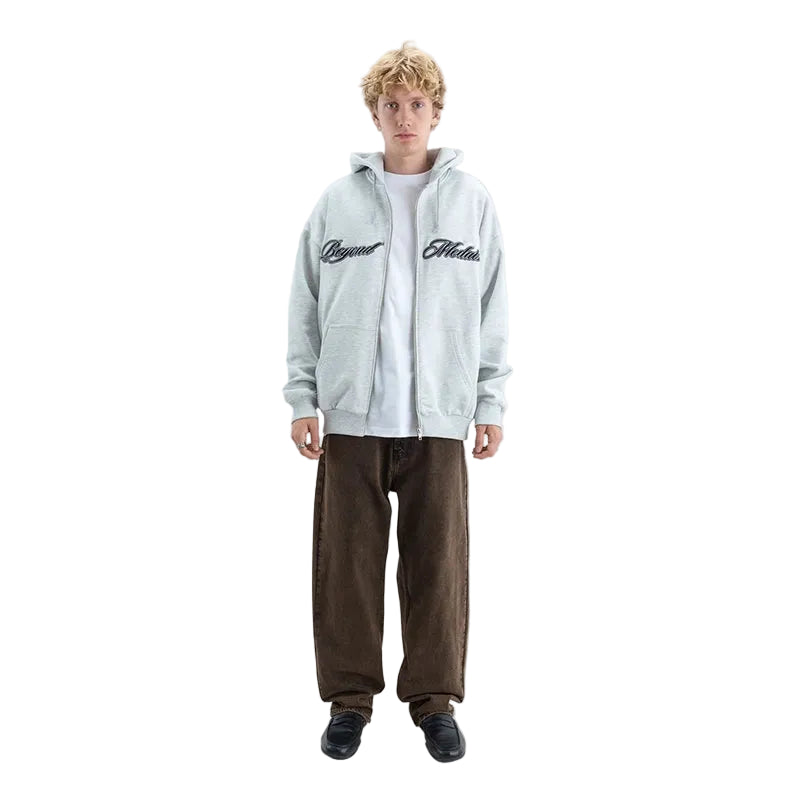A model stands against a light gray studio background wearing a light gray zip-up hoodie with black script embroidery on the chest, a white t-shirt, brown corduroy pants, and black shoes.