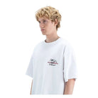 A young man with curly blonde hair wearing an oversized white t-shirt with a small graphic on the left chest, photographed against a neutral light gray background.