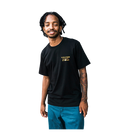 A man wearing a black Volcom Stone t-shirt with a gold logo on the left chest, paired with teal pants, isolated on a white background.