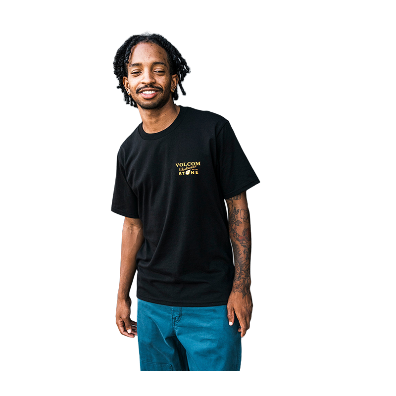 A man wearing a black Volcom Stone t-shirt with a gold logo on the left chest, paired with teal pants, isolated on a white background.