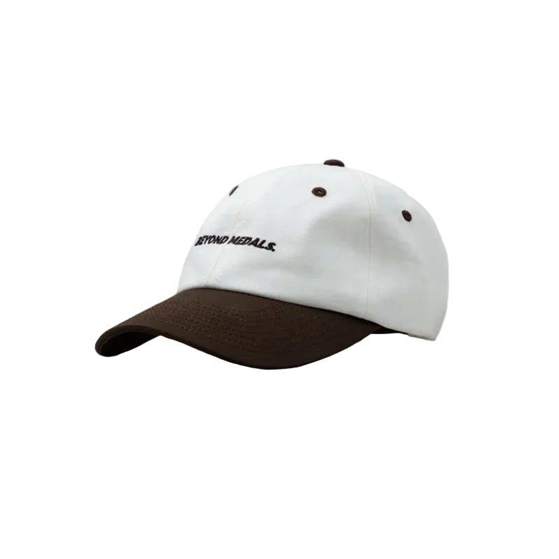 A baseball cap with a white crown and a dark brown visor, featuring the embroidered text 'BEYOND MEDALLS' in black on the front panel. The cap is photographed from a three-quarters angle against a solid light gray background.