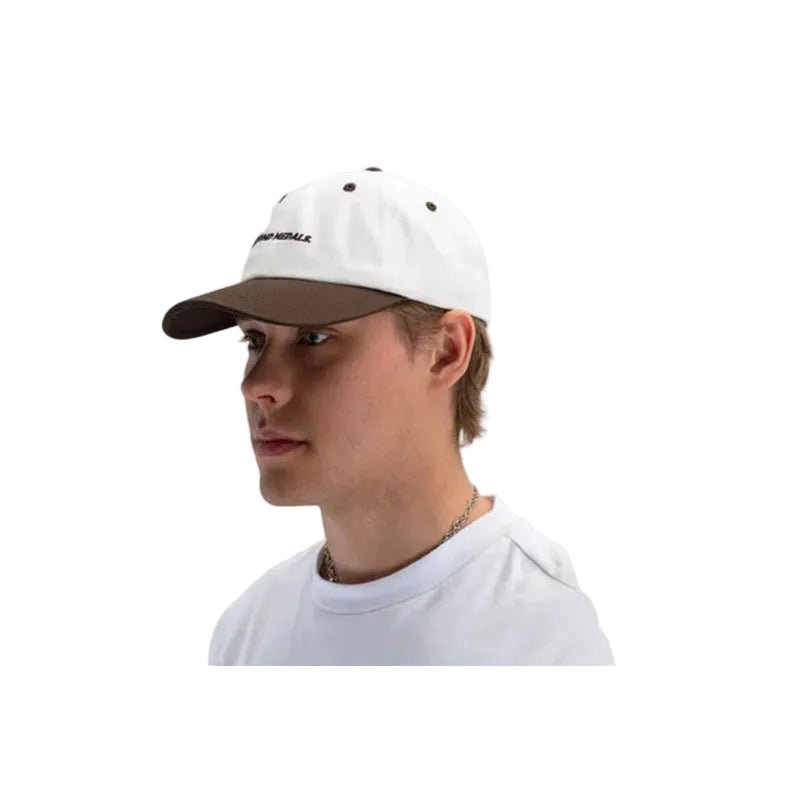 A man wearing a white baseball cap with a brown visor and black embroidered text on the front, paired with a white t-shirt against a light gray background.